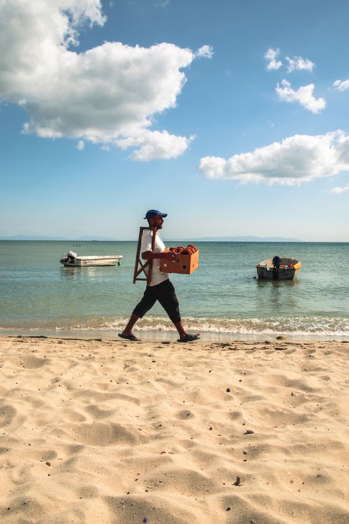 Free A vendor walks along a sunny, serene beach carrying goods, with boats resting on the calm sea. Stock Photo