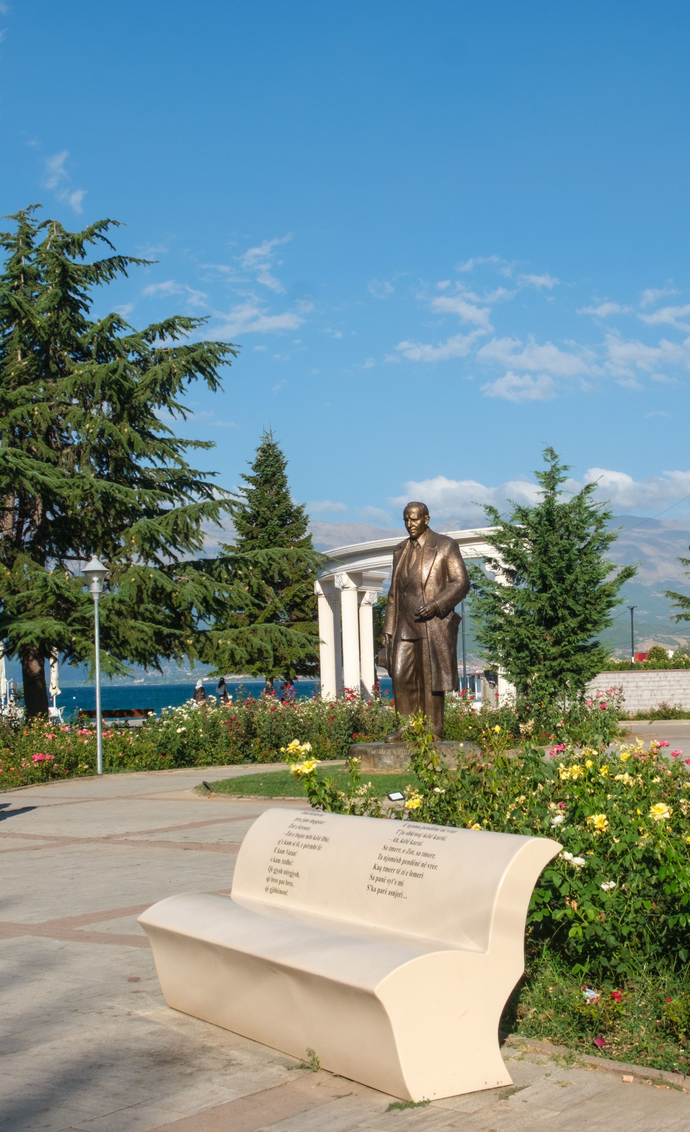 A serene park scene in Pogradec, Albania featuring a prominent statue surrounded by flowers under a bright sky.