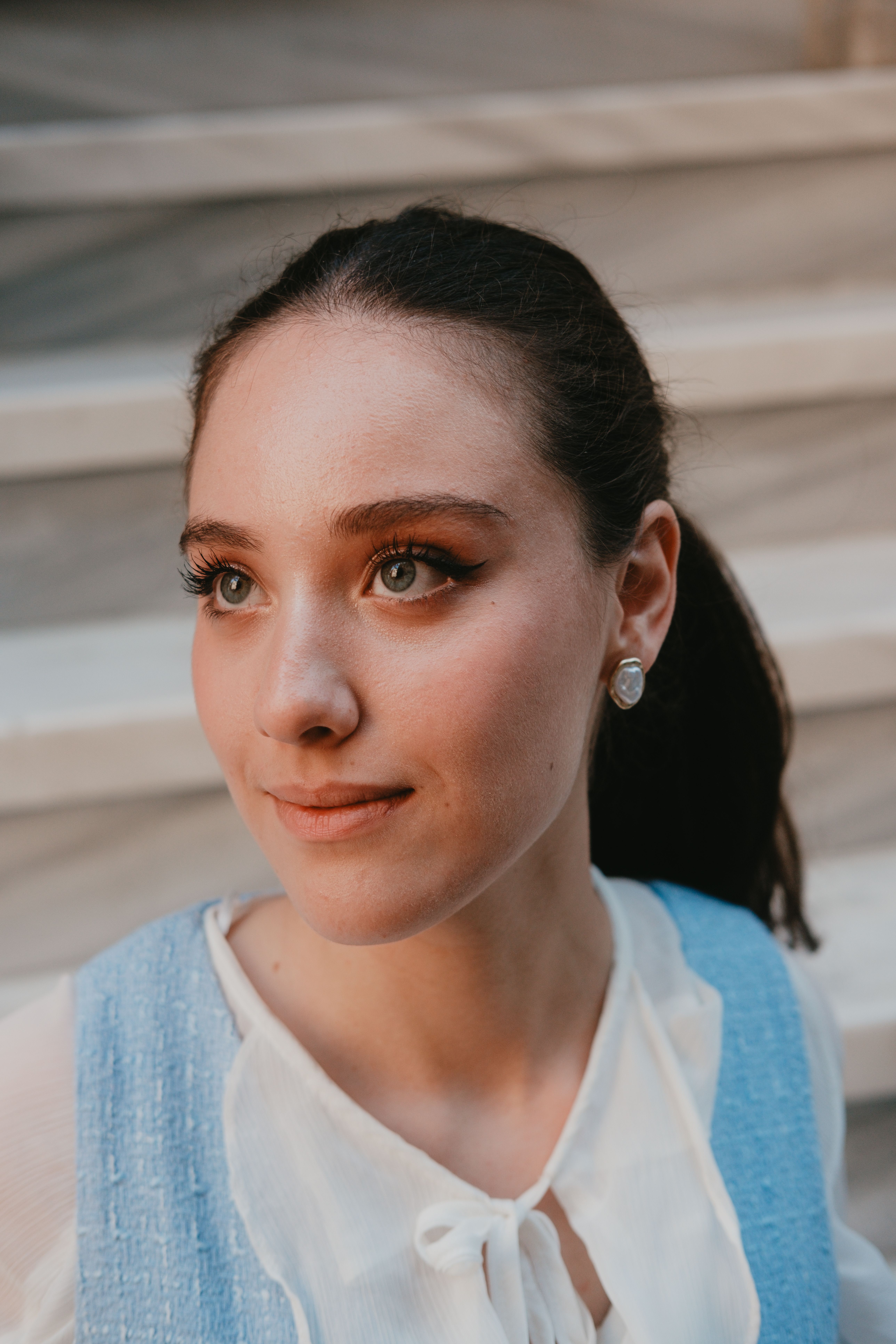 Portrait of a young woman with soft expression in elegant blue and white outfit, sitting on marble steps.