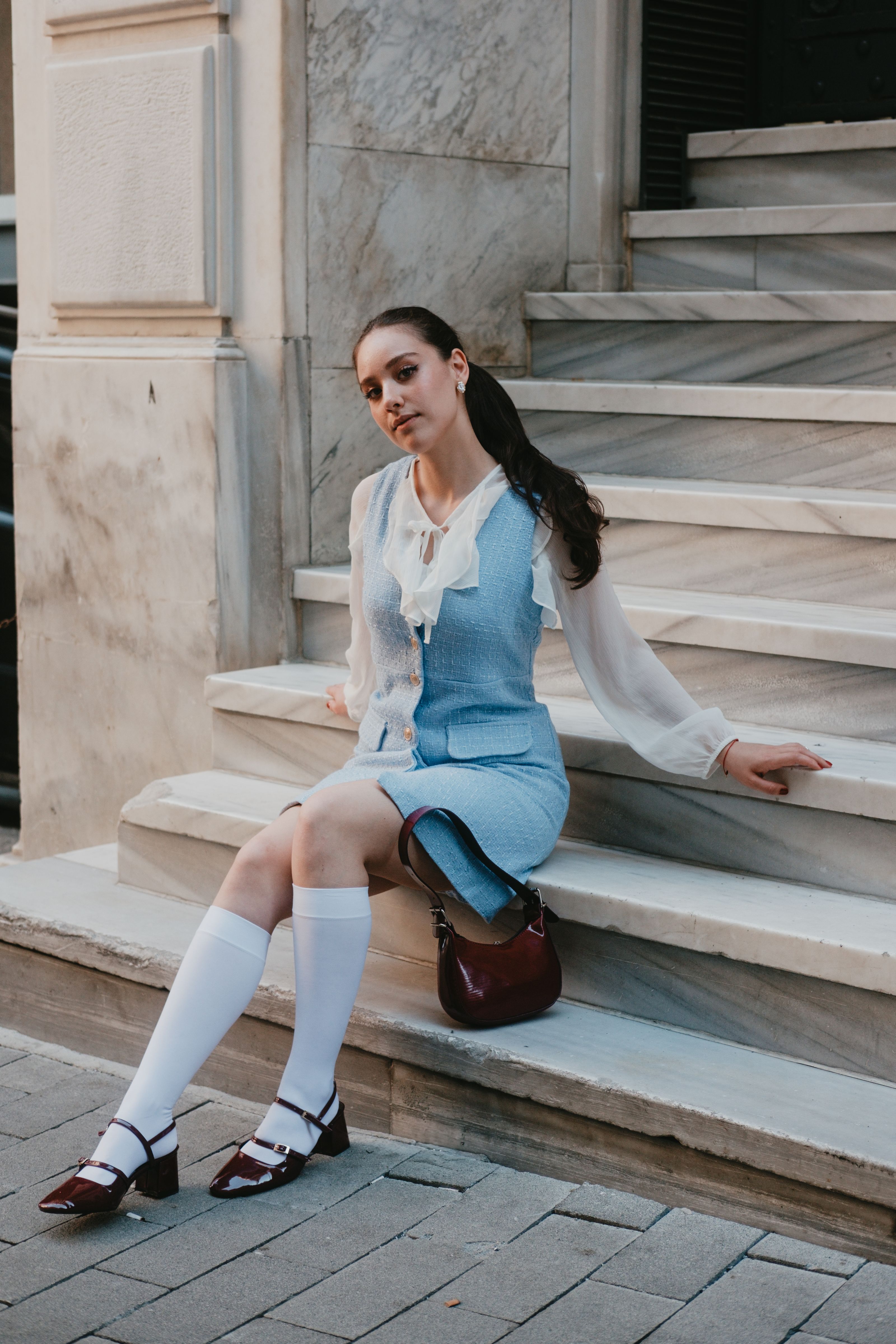 Uma jovem mulher de vestido azul e blusa branca está sentada em elegantes degraus de mármore.