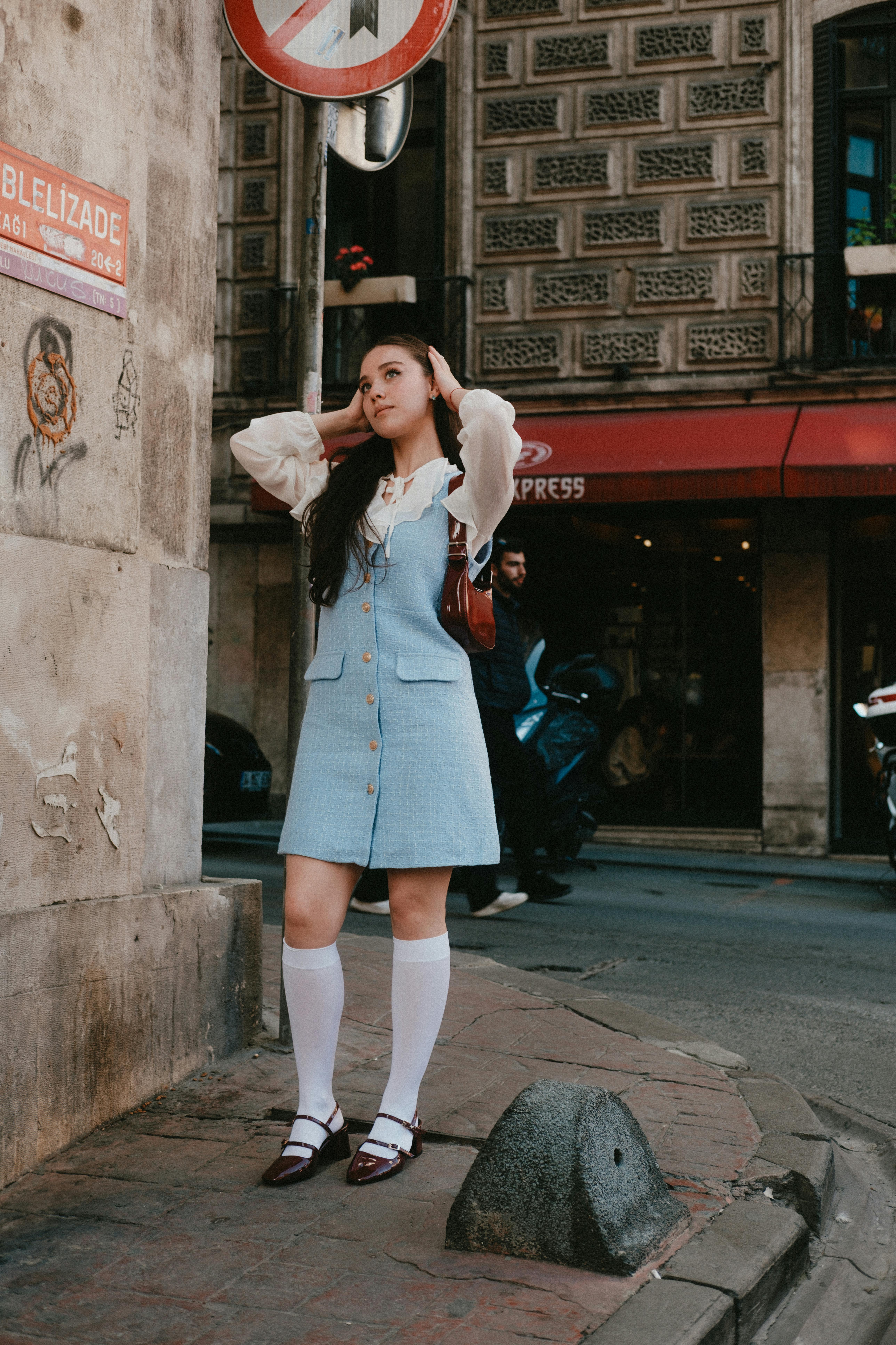 Young woman in stylish outfit poses at an urban street corner with vintage architecture.
