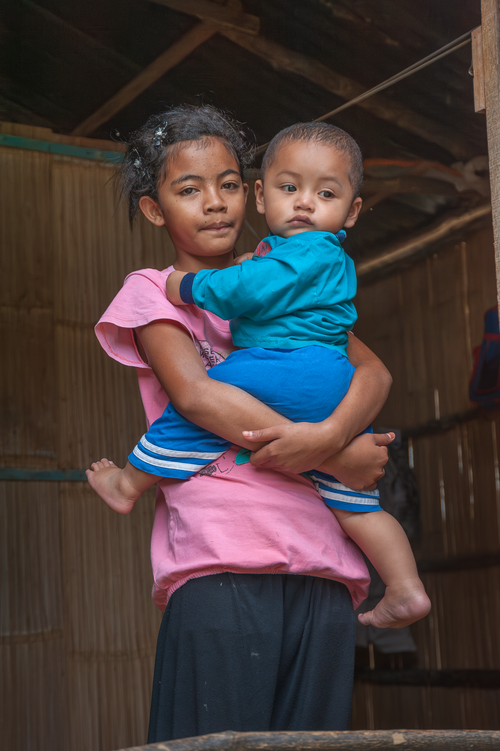 Free Young girl holding her baby sibling, depicting warmth and care in a rustic home. Stock Photo