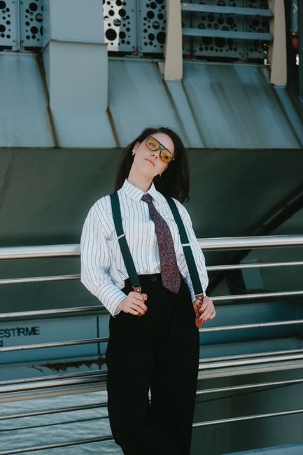 Fashionable woman poses in urban setting wearing suspenders and tie, exuding confidence.