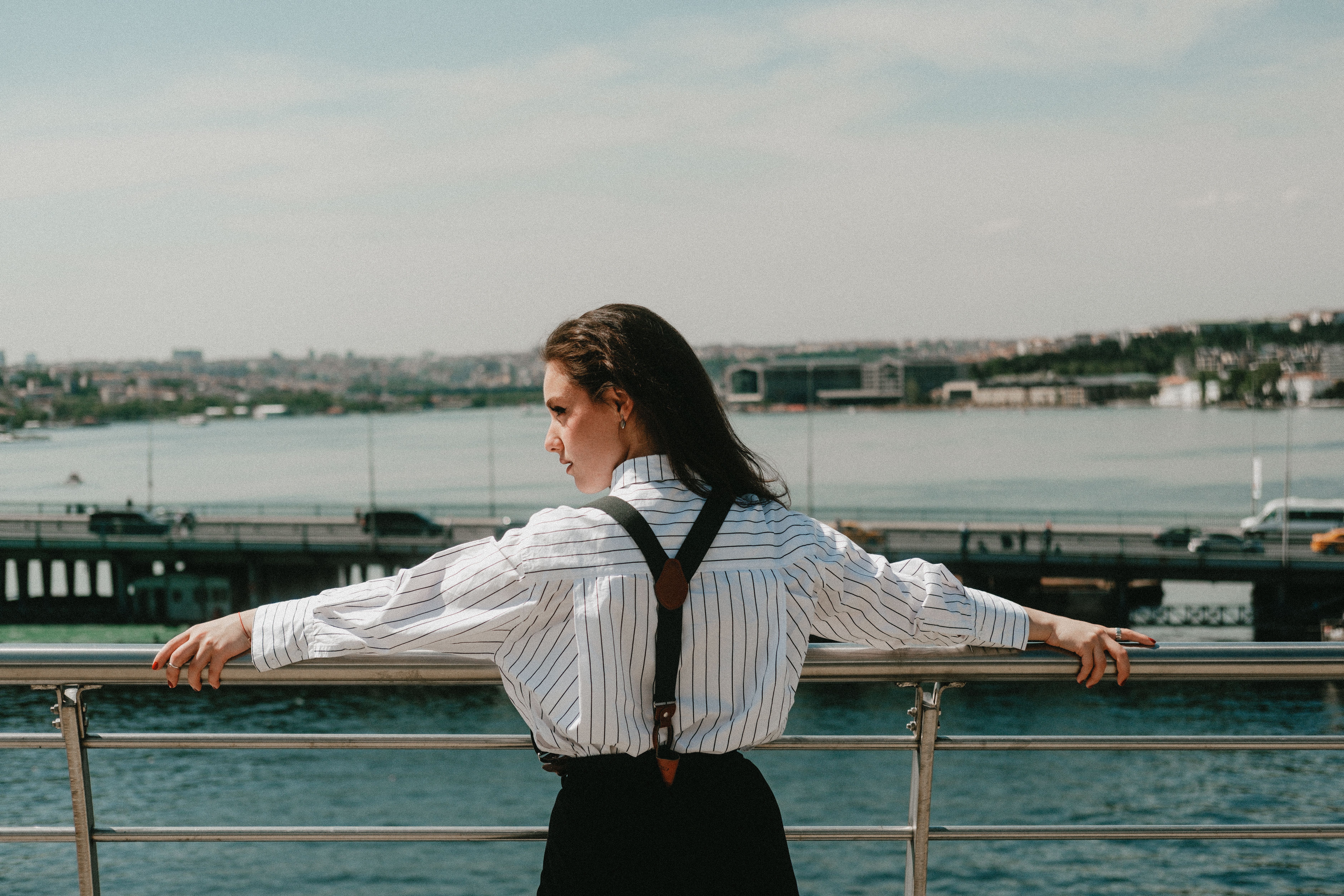 A woman stands on a riverside balcony, gazing at the scenic city landscape under a bright sky.