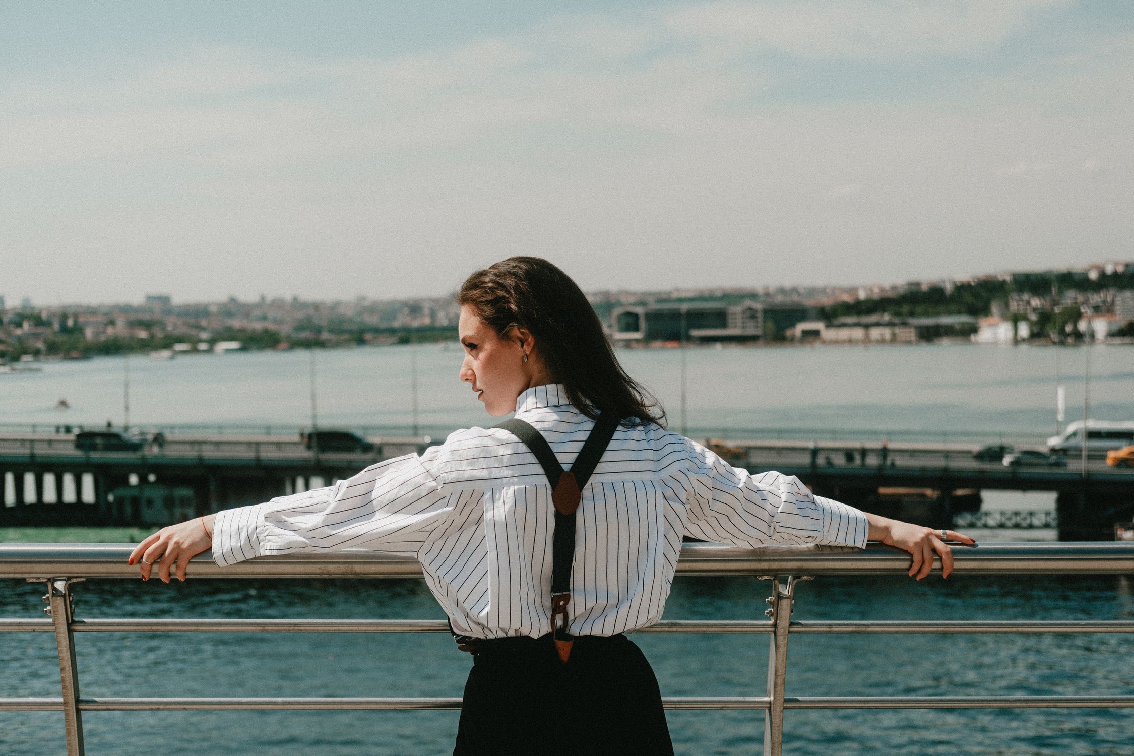 A woman stands on a riverside balcony, gazing at the scenic city landscape under a bright sky.