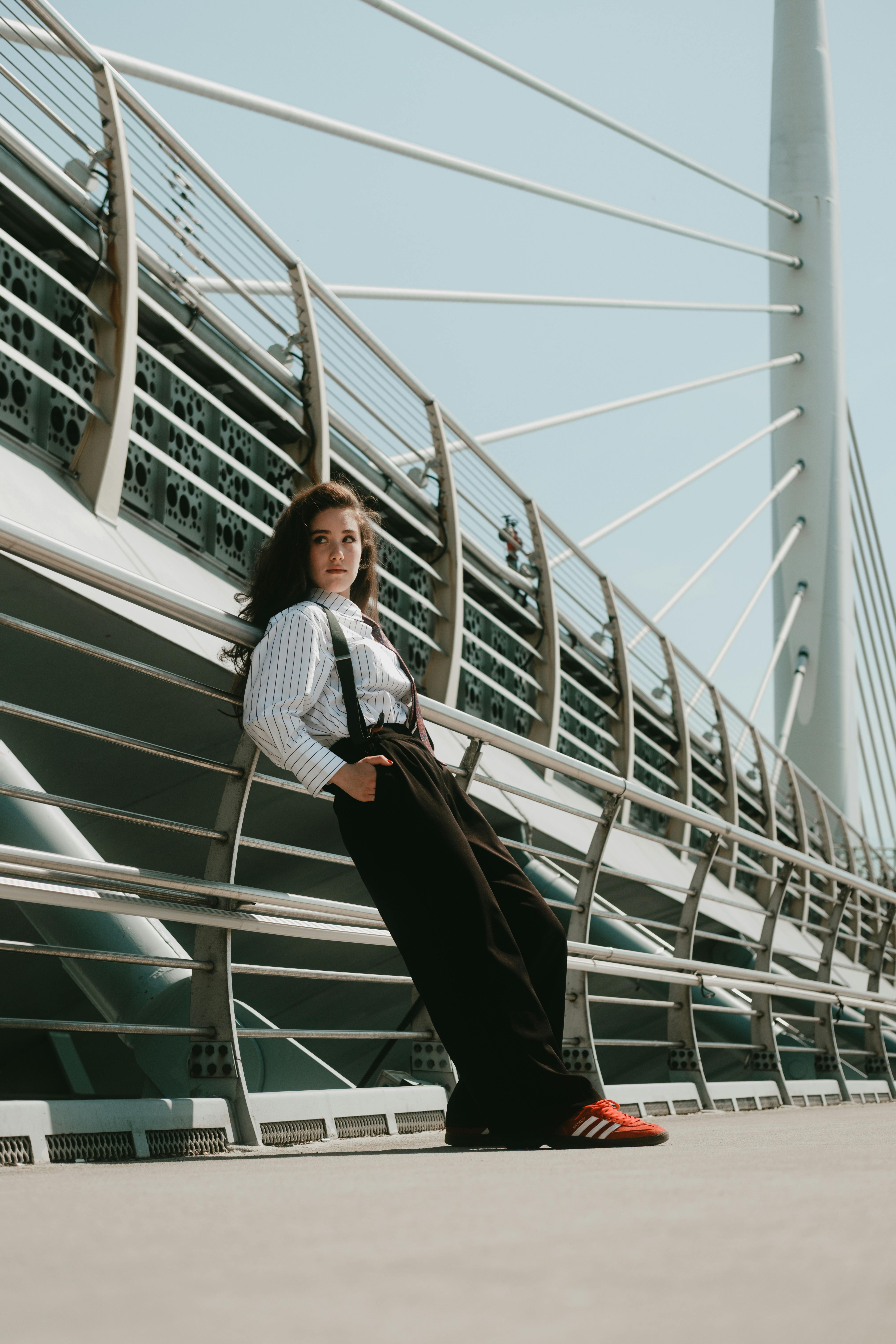 Woman in fashionable attire leaning on a futuristic bridge railing, sunny day.
