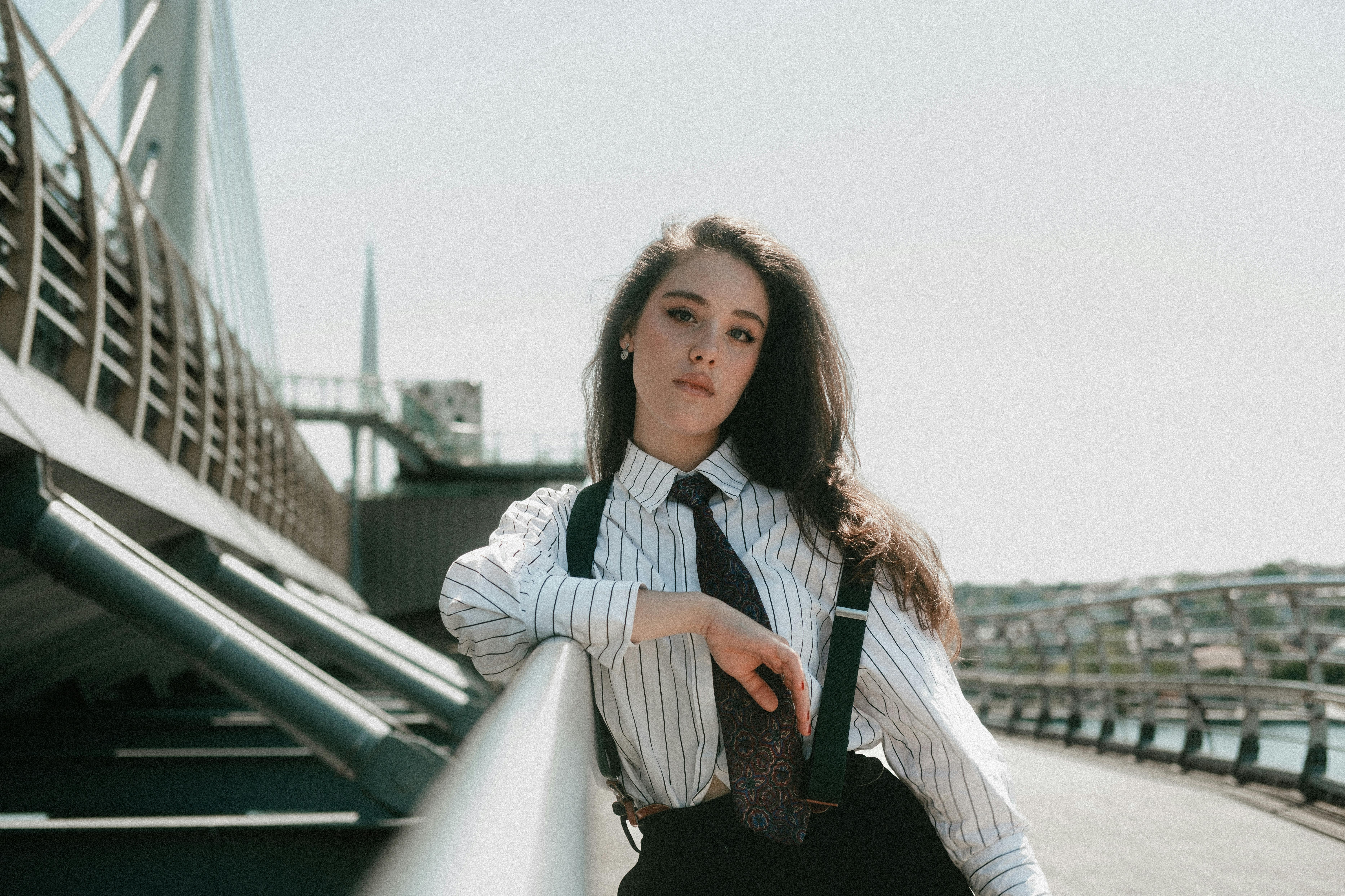 Confident young woman in business attire poses on a modern bridge, embodying urban elegance.