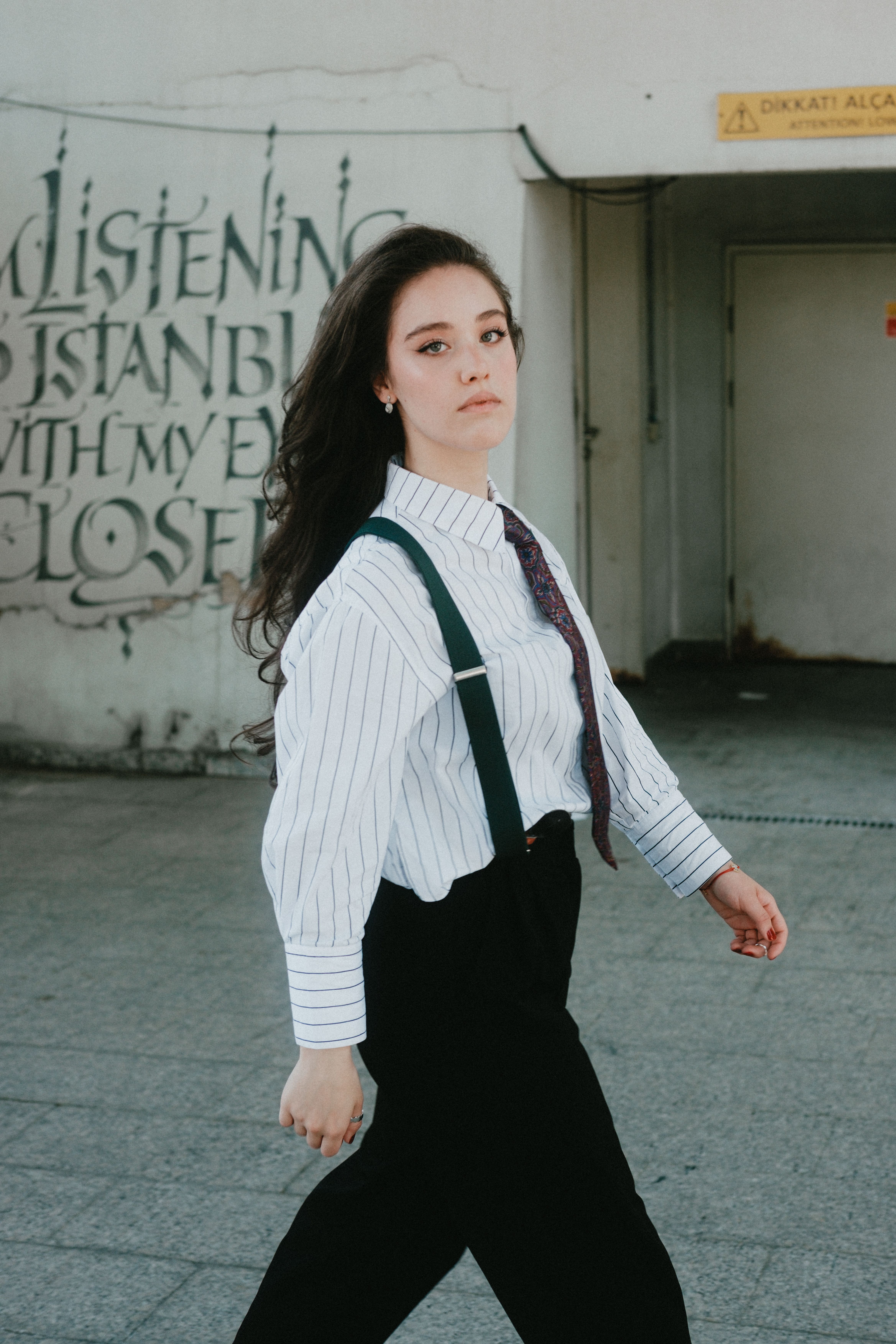 Young woman in striped shirt and suspenders walks past graffiti wall in urban setting.