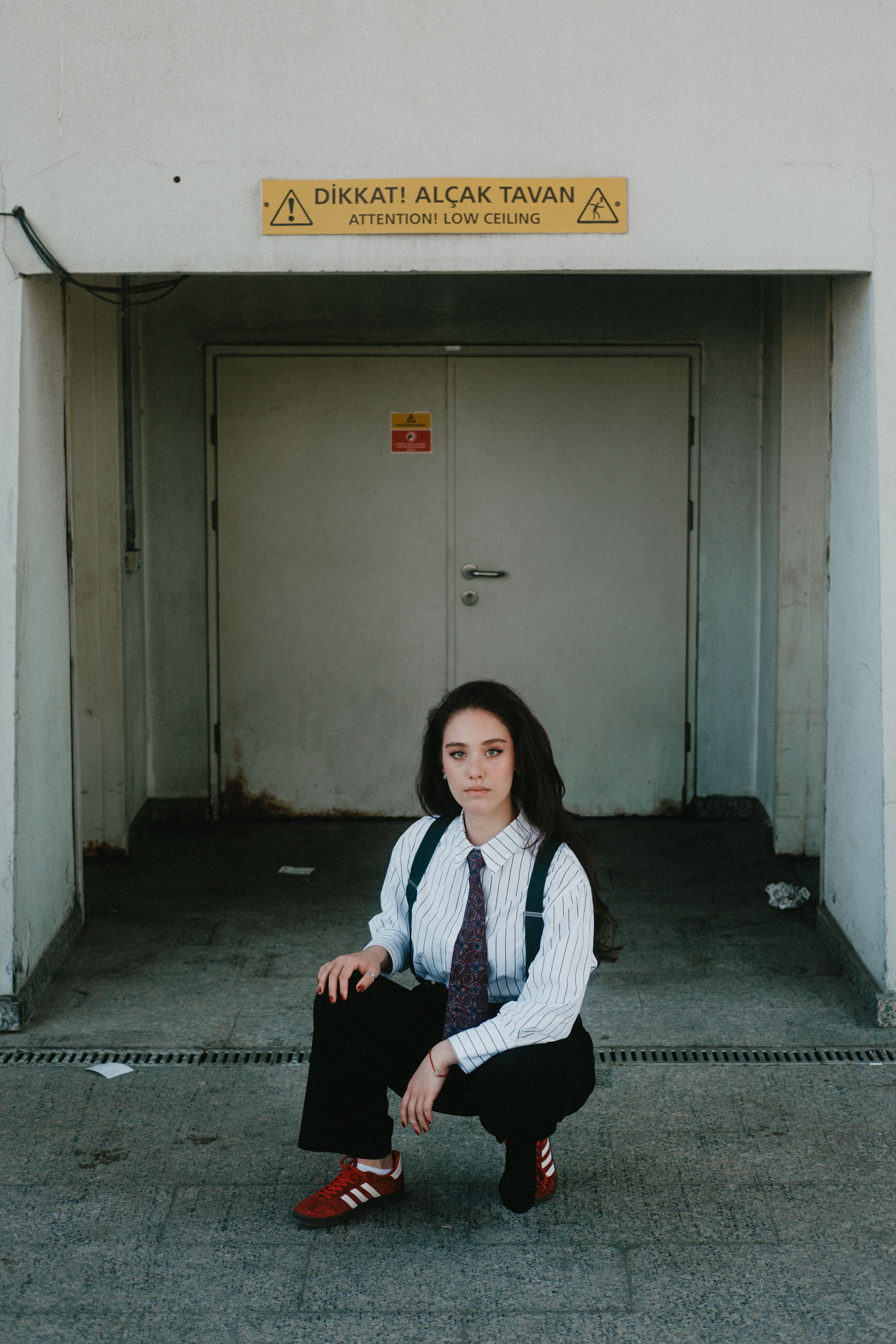 Woman squatting in front of a door with a 'Low Ceiling' warning sign, urban setting.