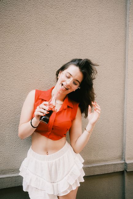 Cheerful woman in red top and white skirt enjoying a soft drink on a sunny day.
