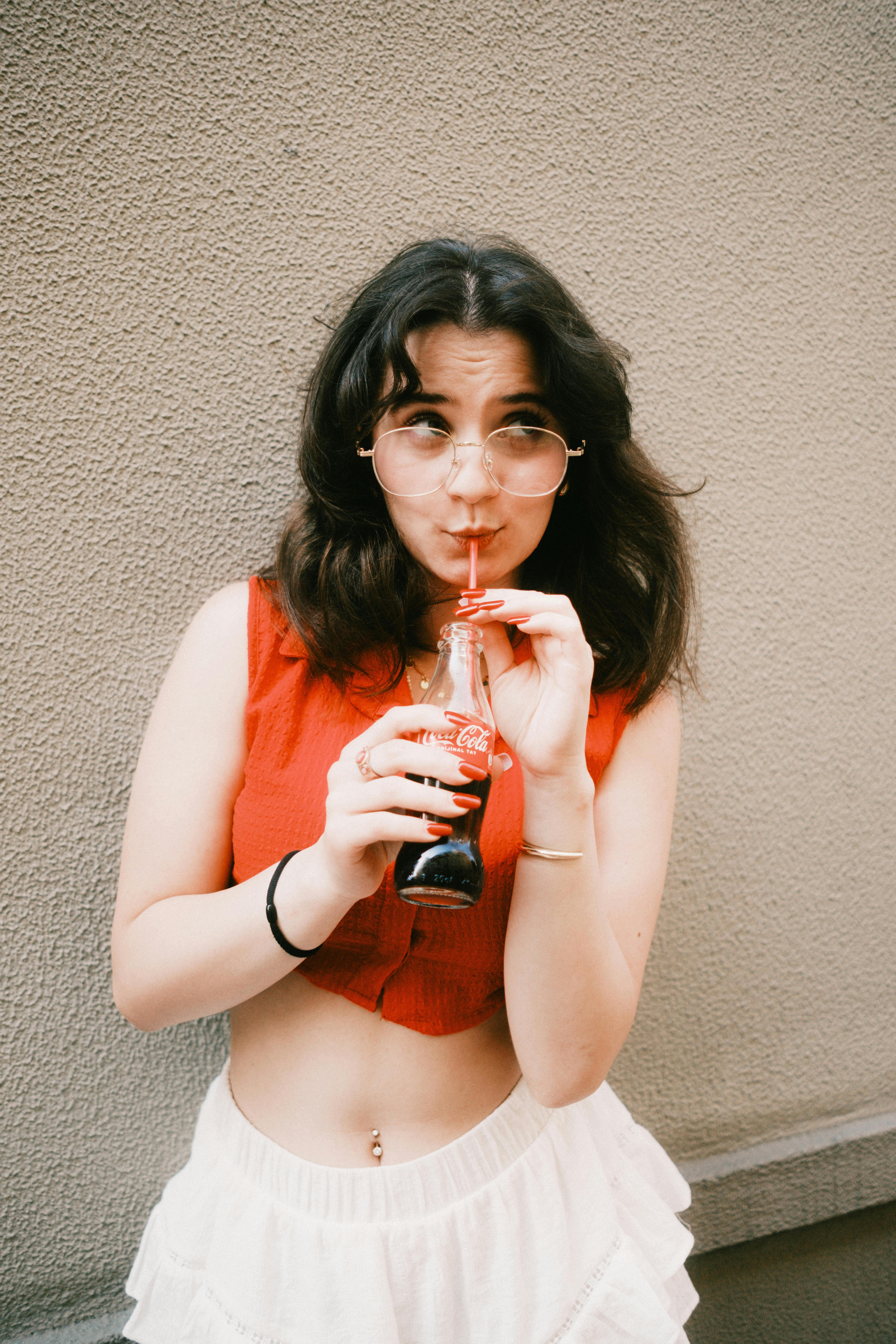 Young woman in red top sipping cola from bottle using a straw against textured wall.