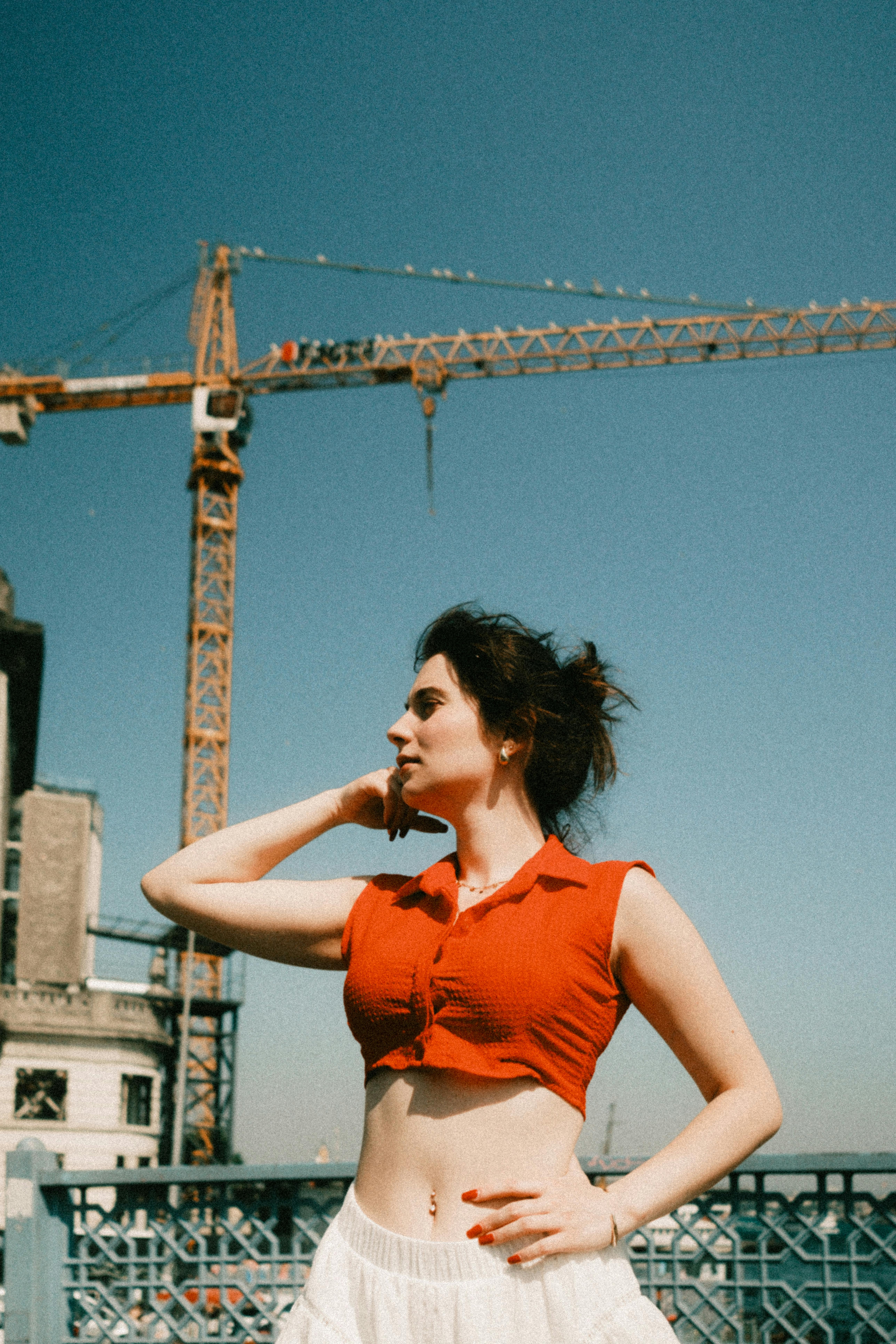 A young woman stands confidently outdoors in İstanbul with a construction crane in the background, under a clear blue sky.