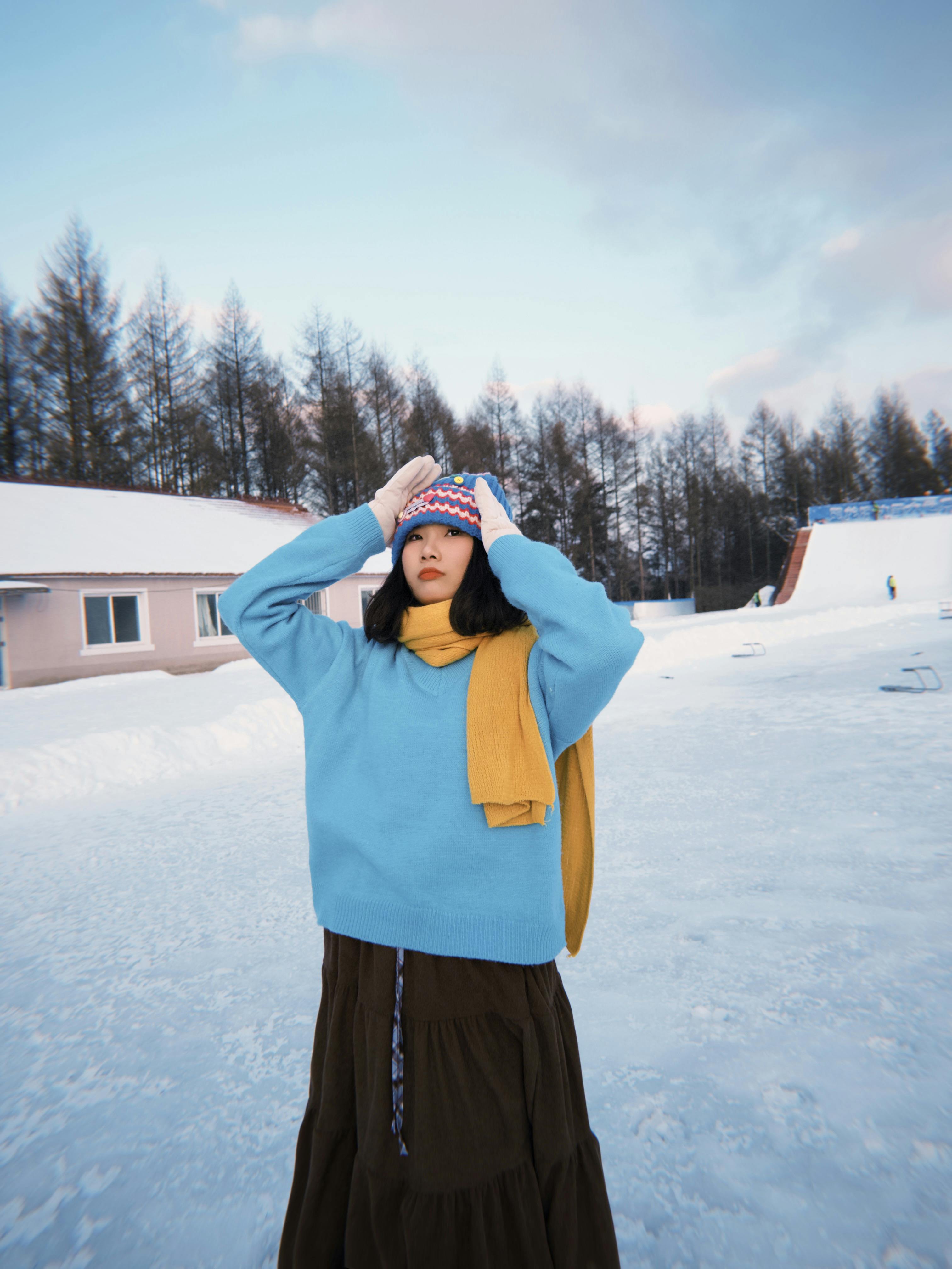 Young woman in winter attire stands in snowy landscape with pine trees.