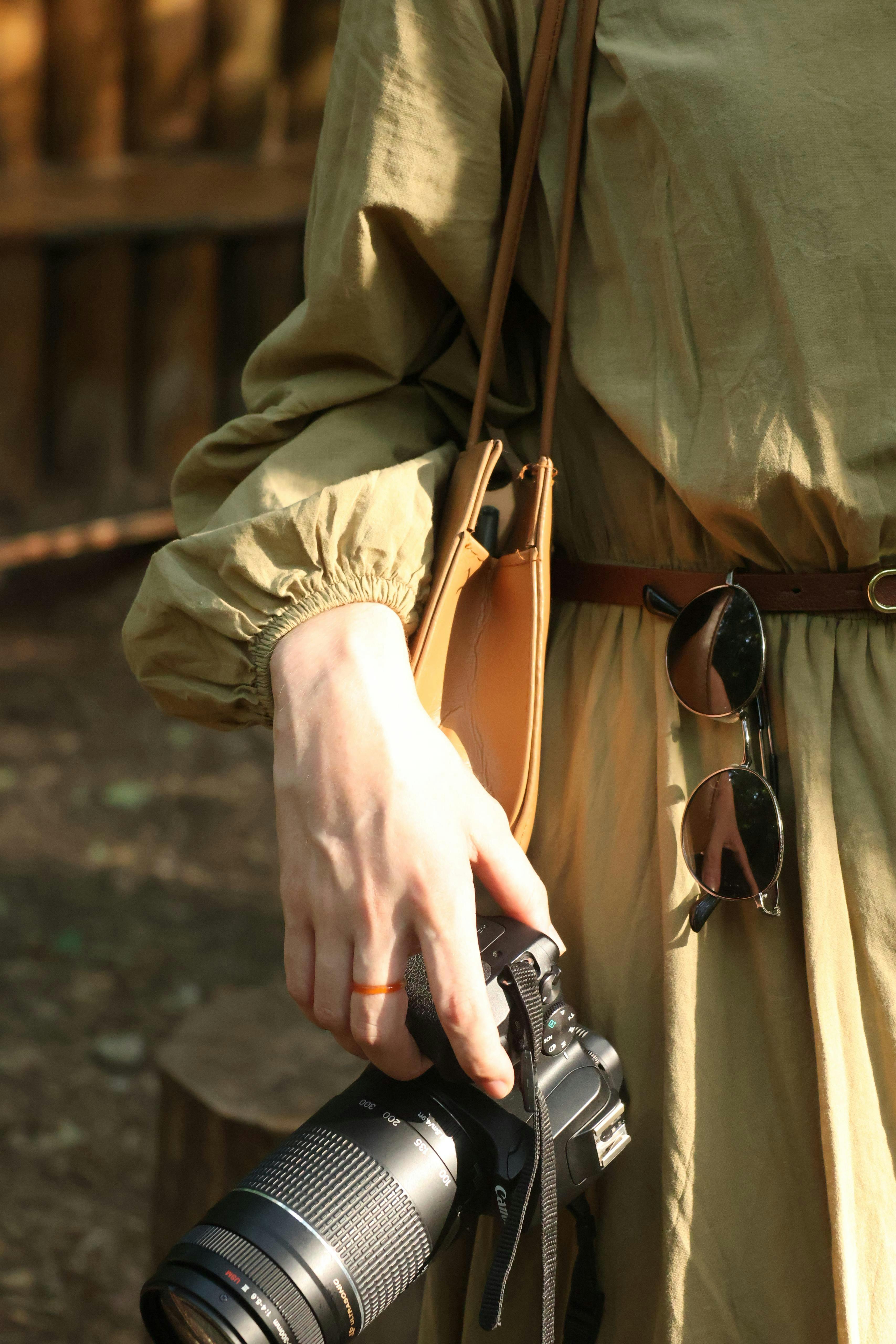A close-up of a person holding a camera outdoors, featuring sunglasses and a stylish outfit.