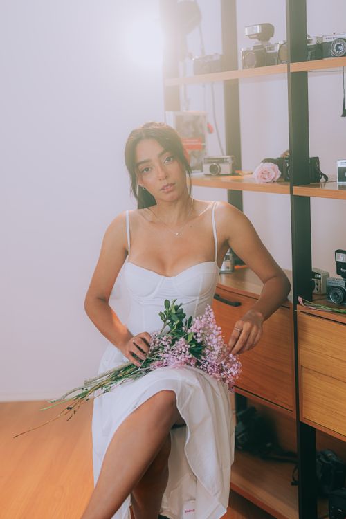 Free Woman in a white dress holding pink flowers in a room with vintage cameras, exuding elegance. Stock Photo