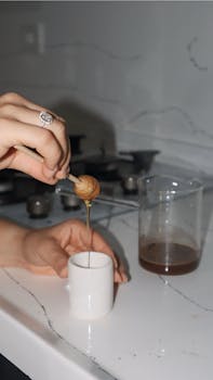 Hand pouring golden honey into a small white cup in a sleek kitchen setting.