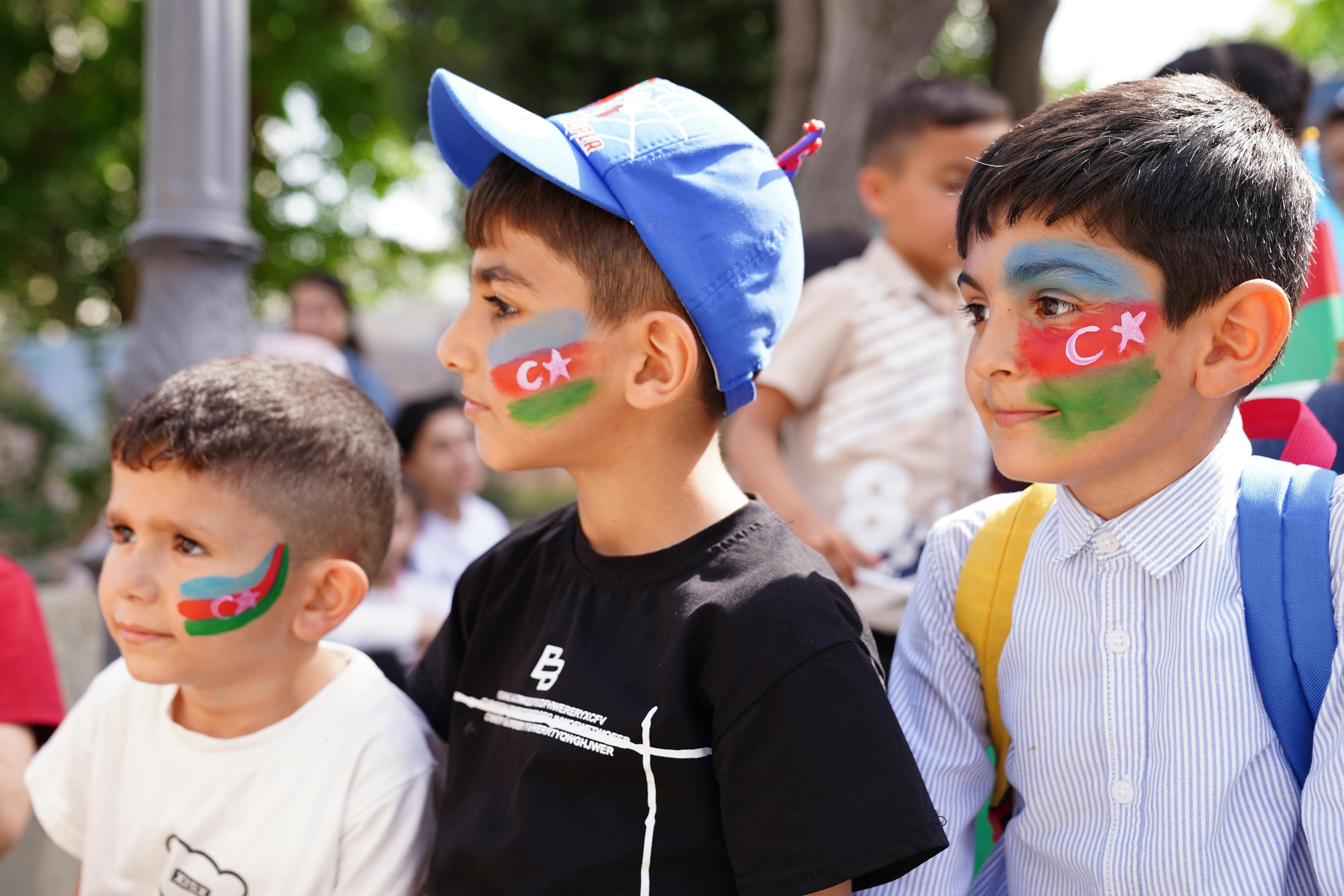 Three boys with Azerbaijan flag face paint smile at an outdoor event. Vibrant and lively scene.