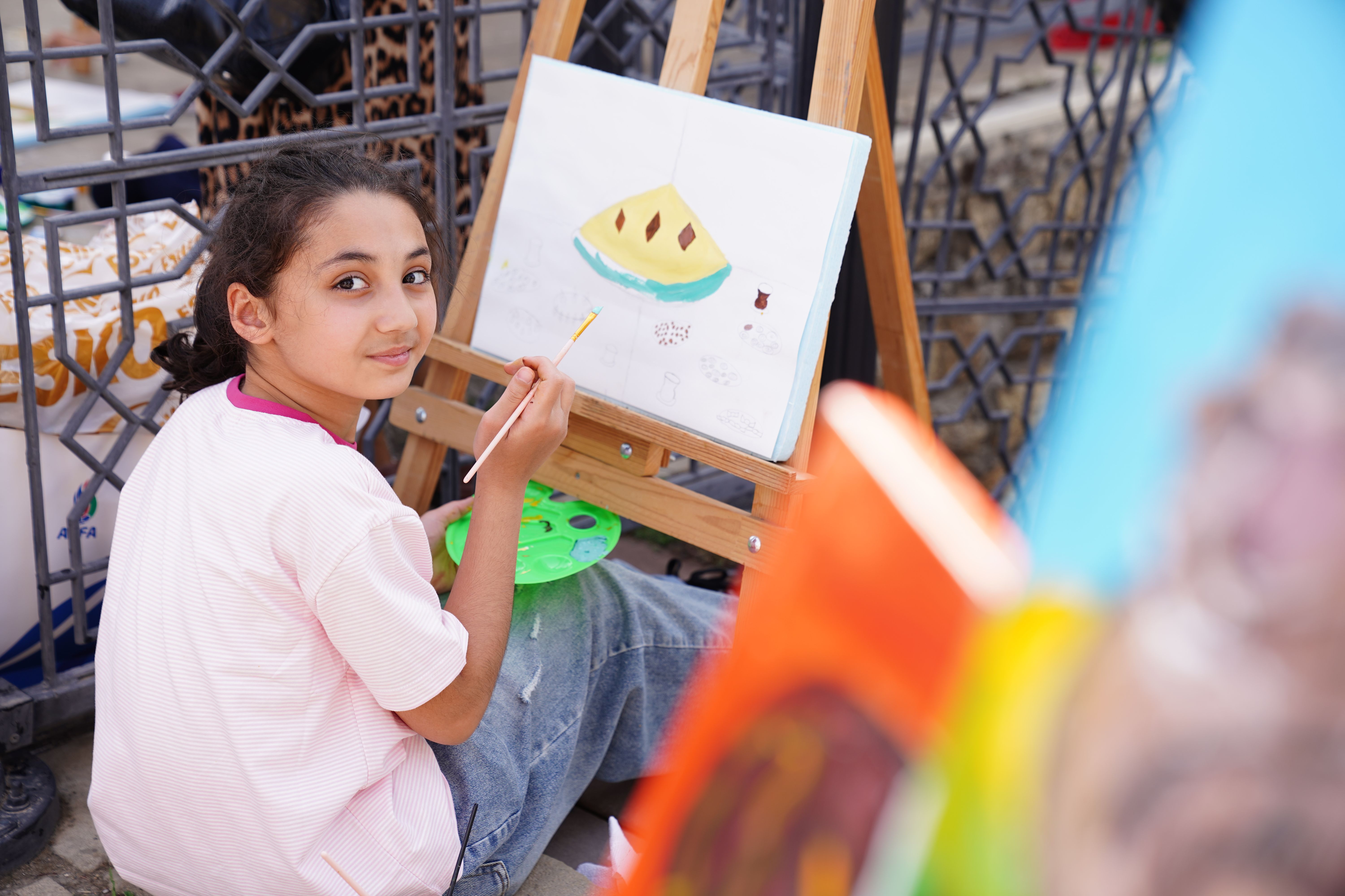 A young girl painting on an easel at an outdoor art event, expressing creativity and joy.