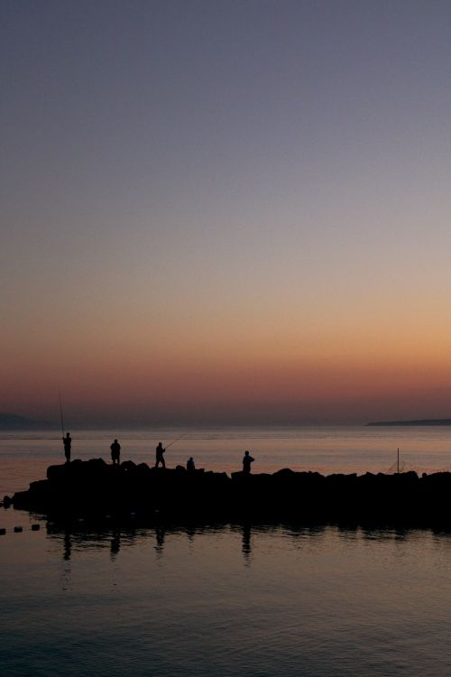 Free Vibrant twilight captures silhouetted fishermen on a rocky pier with calm waters. Stock Photo