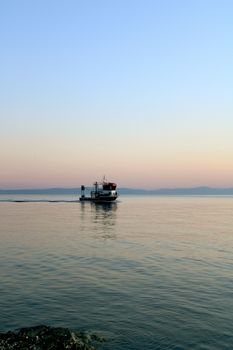 A boat sails at sunset on a calm sea with a pastel sky, creating a peaceful scene.