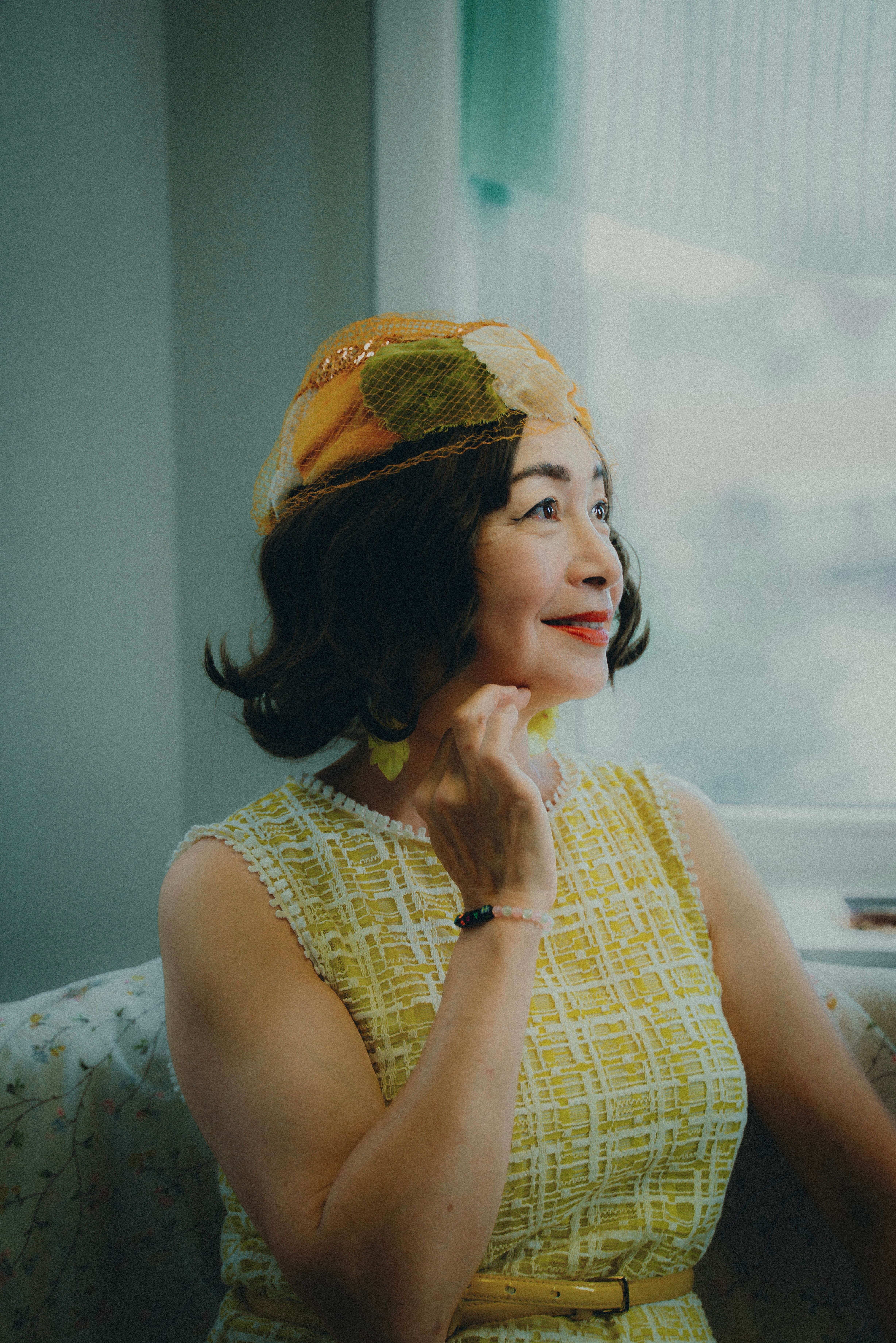 Portrait of a stylish woman in a yellow vintage dress and hat, sitting by a window with natural light.