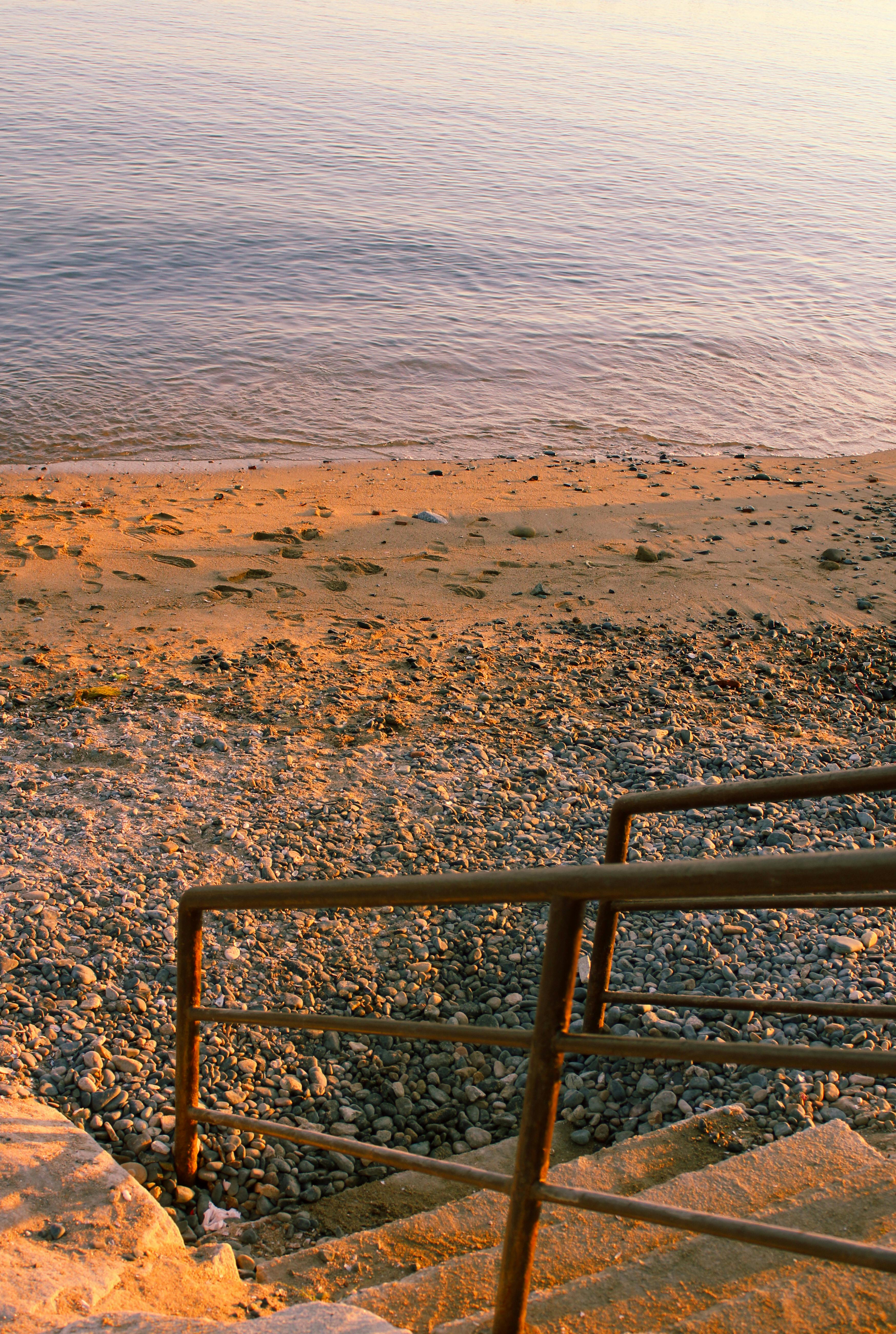 Serene view of a pebbled beach with a staircase descending towards the calm sea under a warm sunset.
