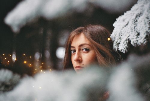 Free A woman peeks through snow-covered branches, her gaze mysterious amidst winter lights. Stock Photo