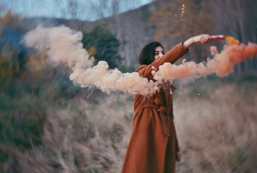 Free Young woman holds an orange smoke bomb in a misty, autumnal setting outdoors. Stock Photo