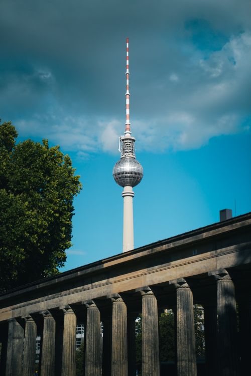 Free The iconic Berlin TV Tower stands tall against a vibrant blue sky, framed by a leafy tree and classical architecture. Stock Photo