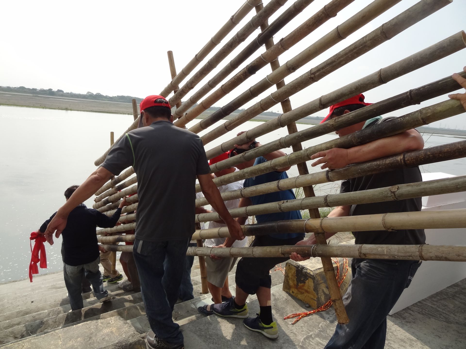 A group of people collaboratively carrying a bamboo structure near a riverside during the day.