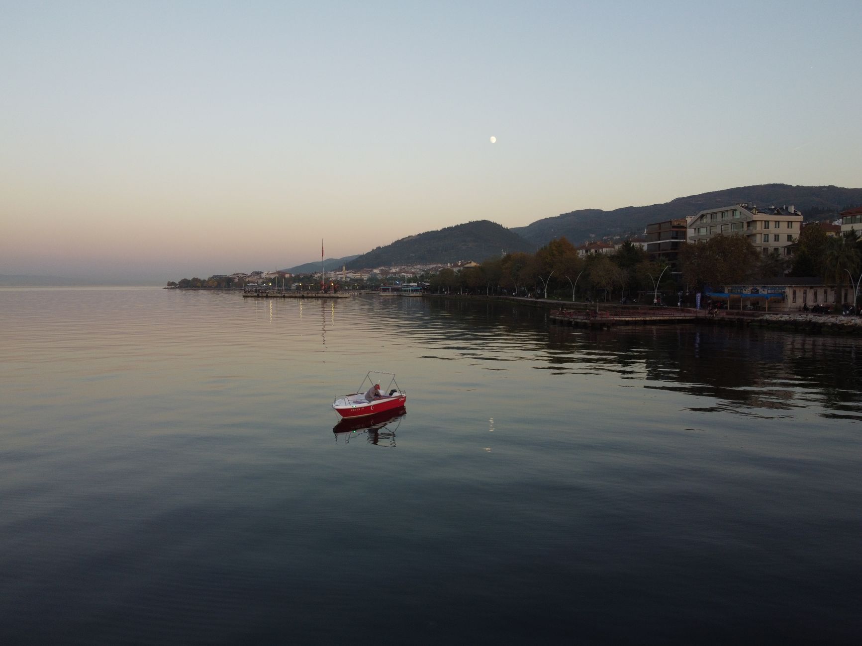 A red boat gently floats on Lake Sapanca under a calm evening sky with the moon.