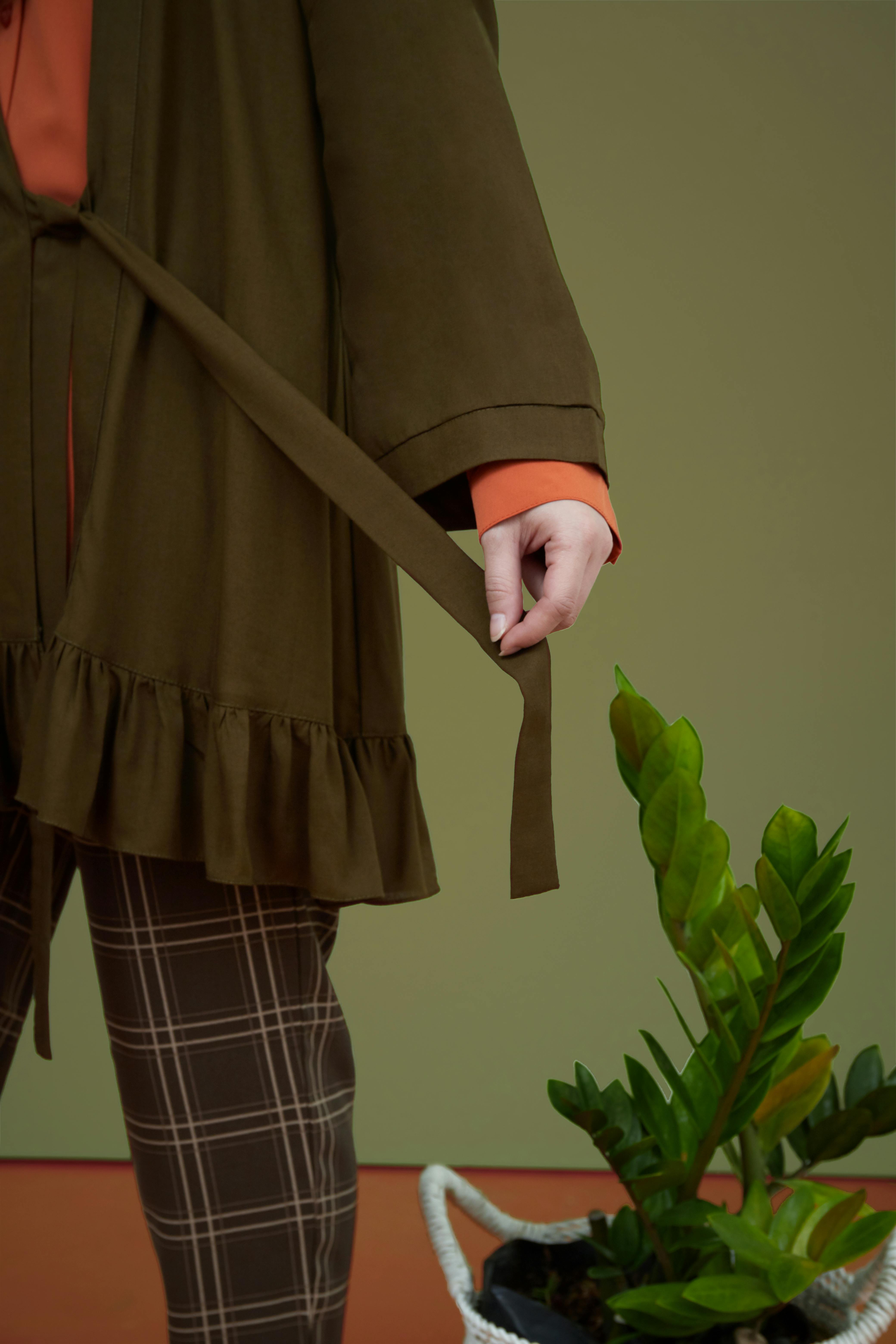 Free Chic fashion ensemble featuring olive green coat, plaid pants, and indoor plant. Stock Photo
