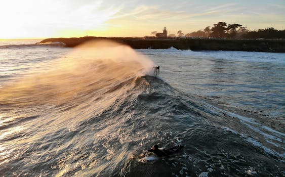 Breathtaking aerial view of surfers riding waves at sunset near the iconic lighthouse in Santa Cruz, California.
