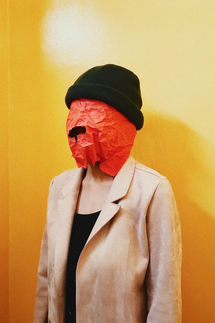 A surreal portrait of a person wearing a red paper mask and green beanie against a yellow background.