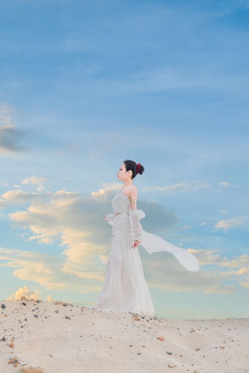 Free Graceful woman in a white flowing dress standing on a sandy hill against a blue sky. Stock Photo