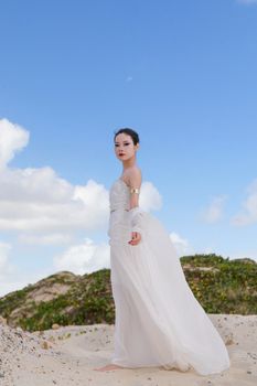 Graceful woman in white dress poses on sandy beach under clear sky.