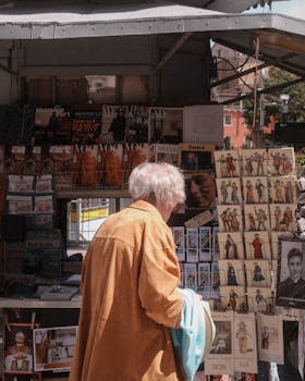 An elderly woman in a beige coat browses vintage magazines at an outdoor kiosk in a sunny European square.