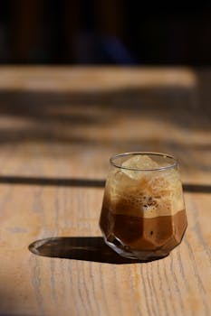 A glass of iced coffee with frothy top on a sunlit wooden table.