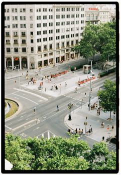 A bustling cityscape with people crossing an intersection, surrounded by greenery and tall buildings.
