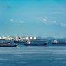 A fleet of oil tankers anchored in calm blue waters near coastal storage facilities.