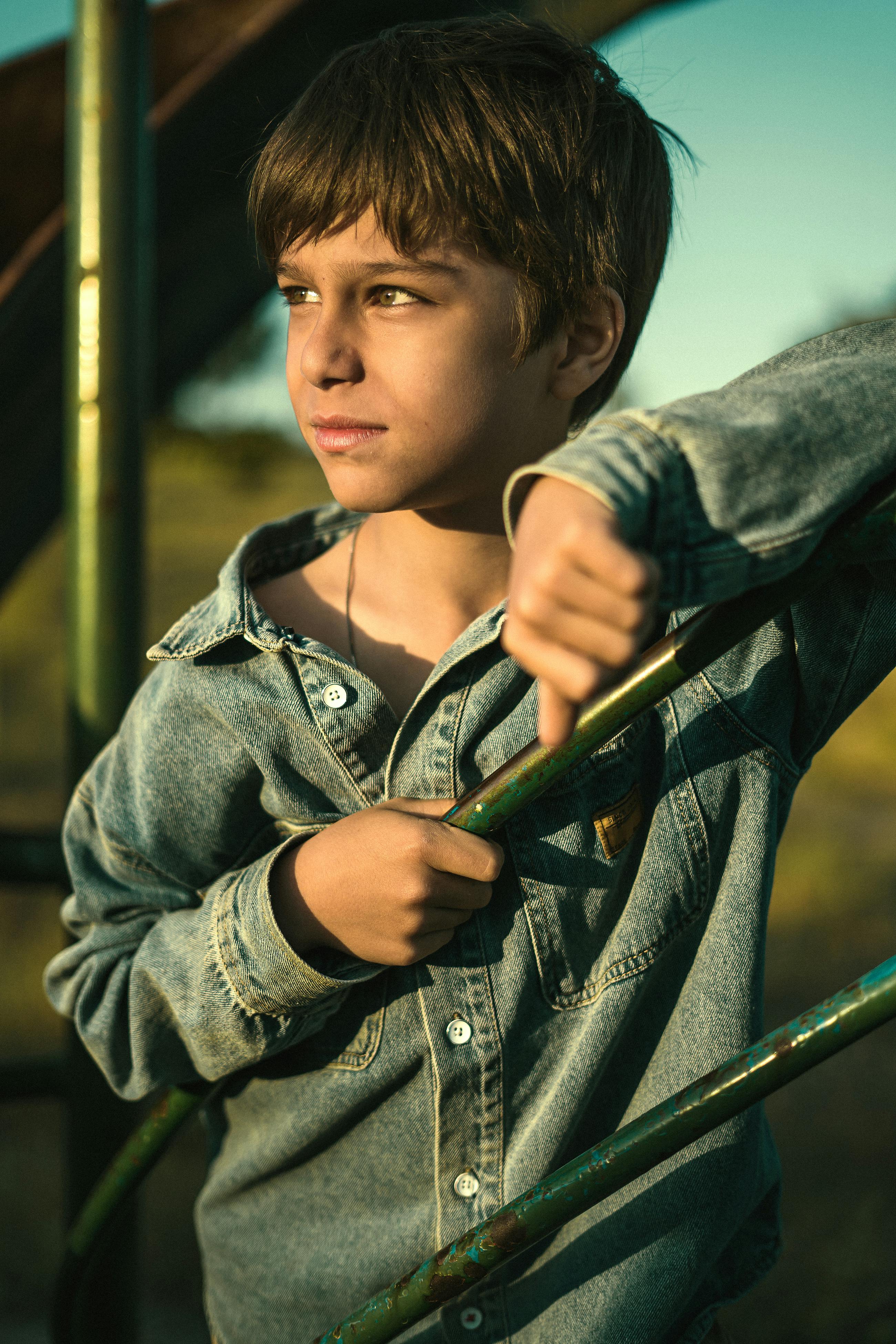A thoughtful young boy in a denim jacket gazes into the distance during sunset, outdoors.