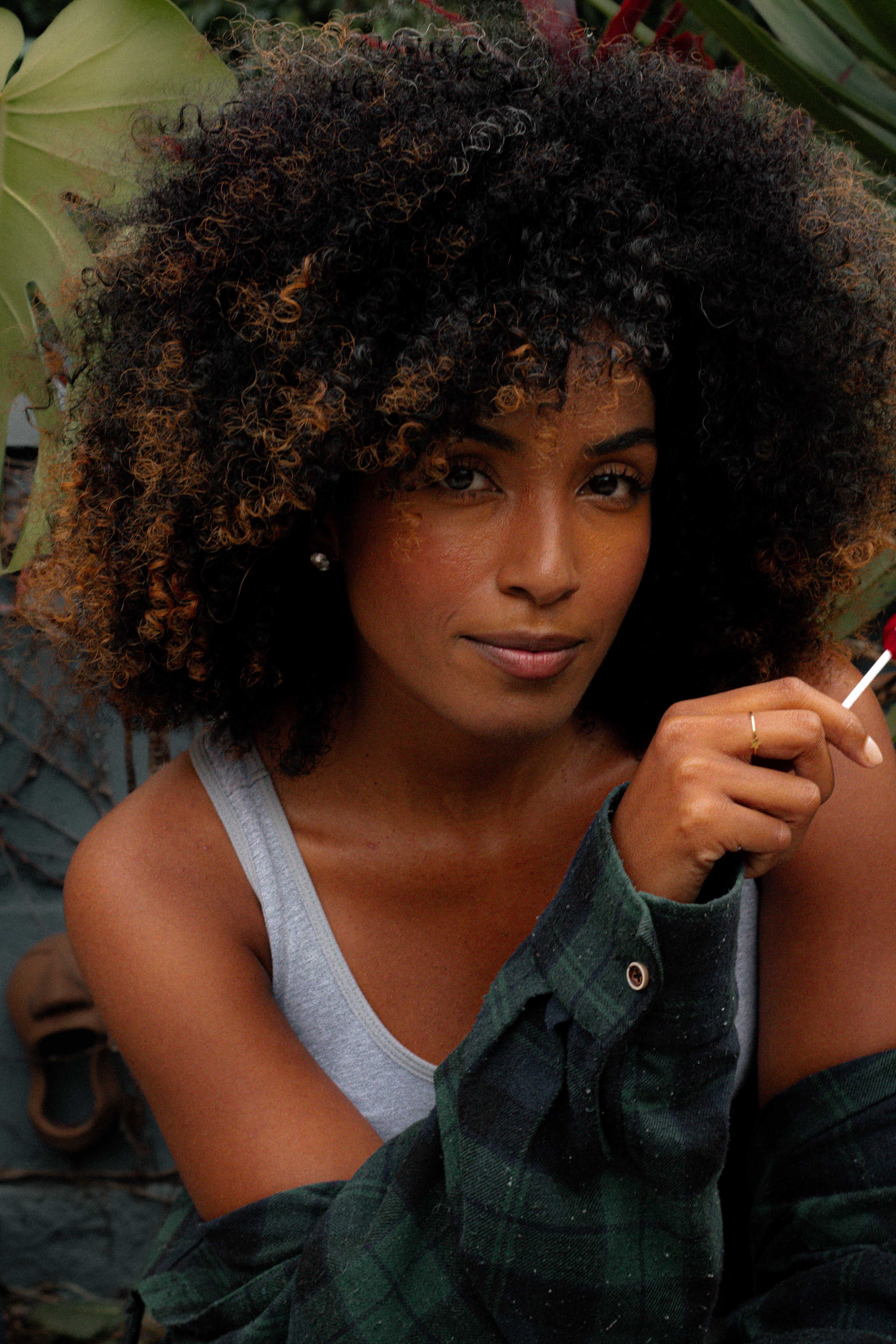 Charming woman with afro hair poses outdoors in casual attire holding a lollipop.