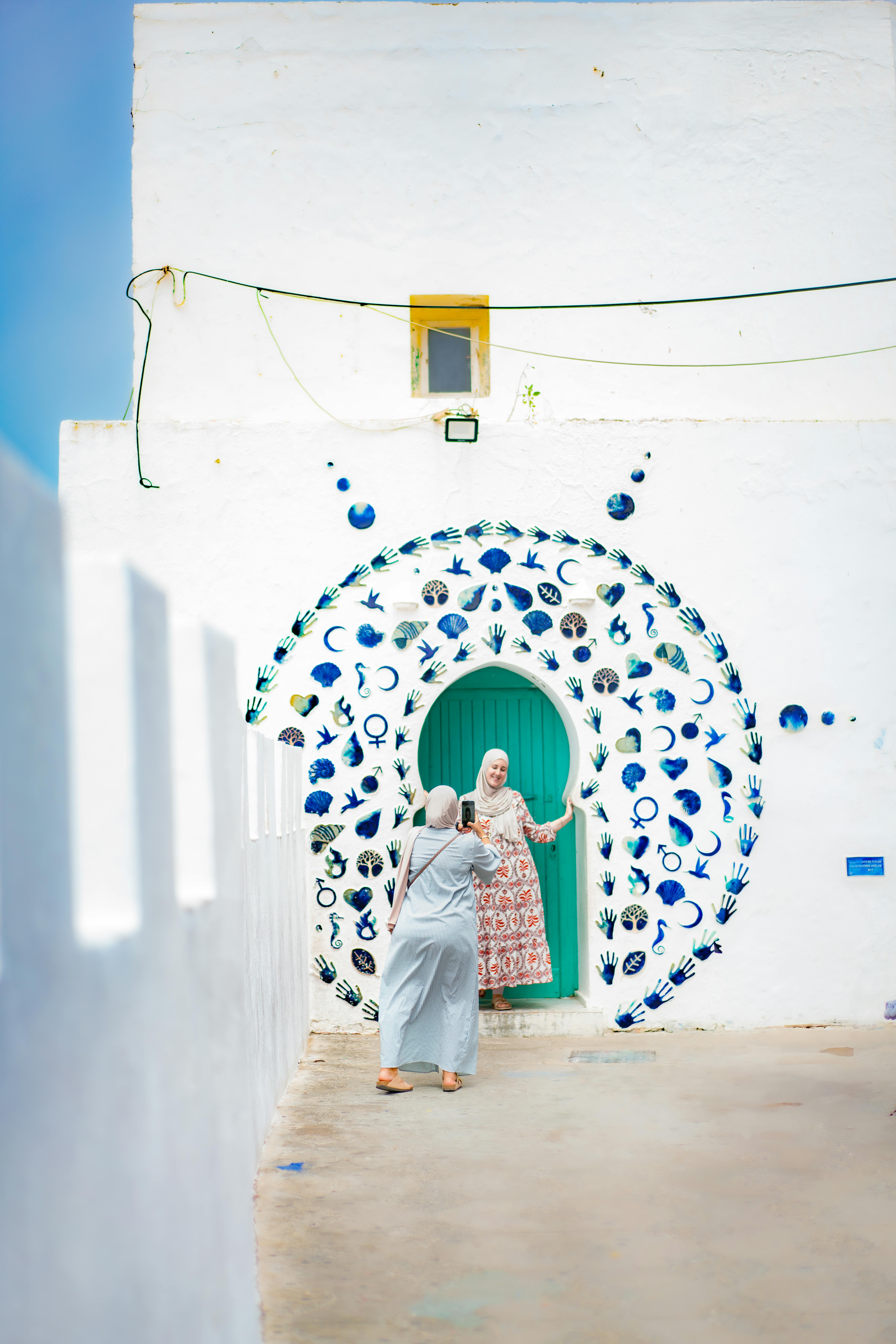 Two people interact near a colorful, artistic wall in a Mediterranean setting.