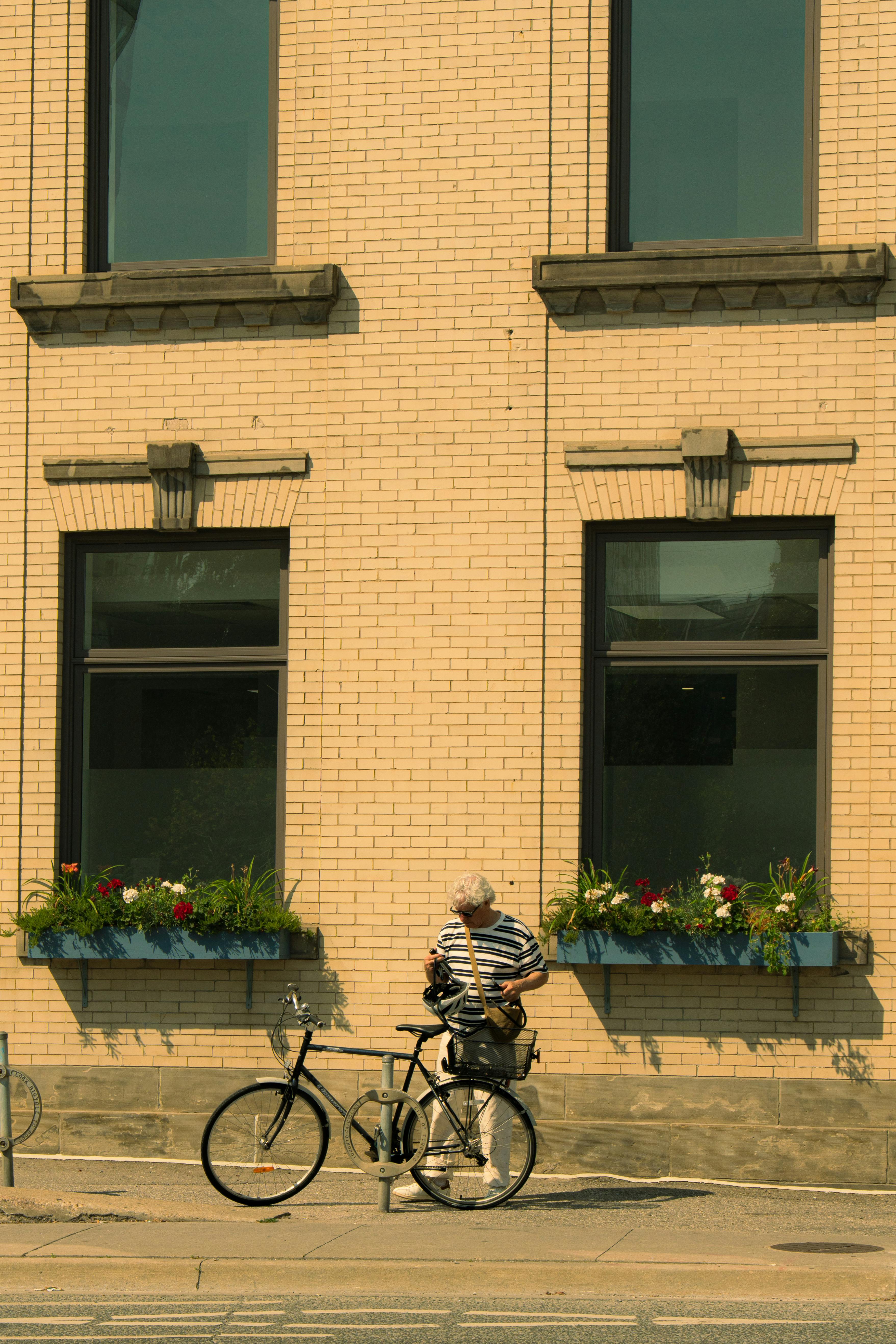 Man with his bike stands by a brick building with flower boxes on a sunny day.