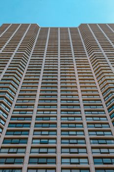A modern high-rise building with numerous windows under a clear blue sky, viewed from below.