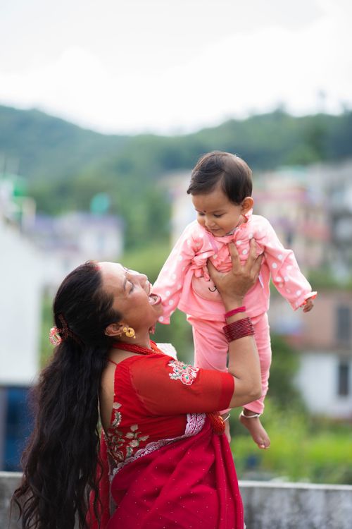 Free Cheerful mother in traditional attire lifts smiling baby outdoors against a scenic backdrop. Stock Photo