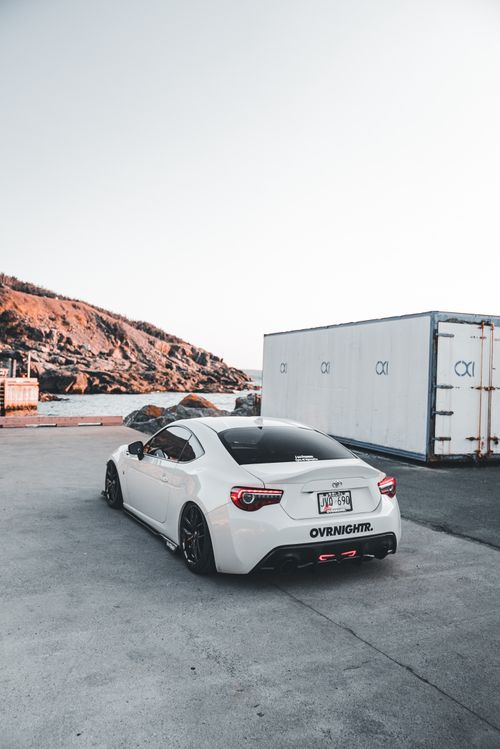 Free White sports car parked near shipping containers by the coast, reflecting modern automotive elegance. Stock Photo