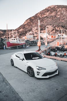 Modern white car parked at a scenic coastal harbor with fishing boats and mountains in the background.