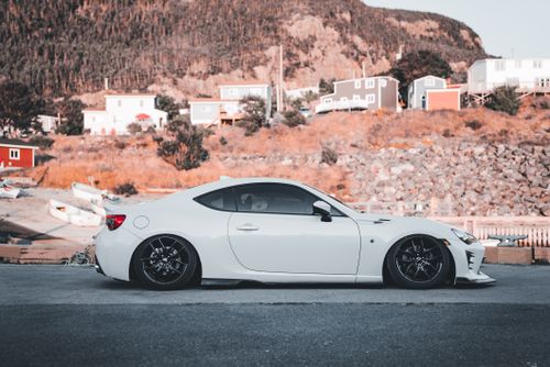 Free White sports car parked in a coastal village setting with scenic mountain background. Stock Photo