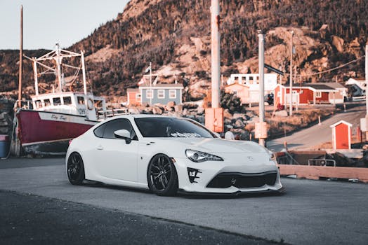 A sleek white sports car parked near a coastal fishing harbor with vibrant boats and colorful houses.
